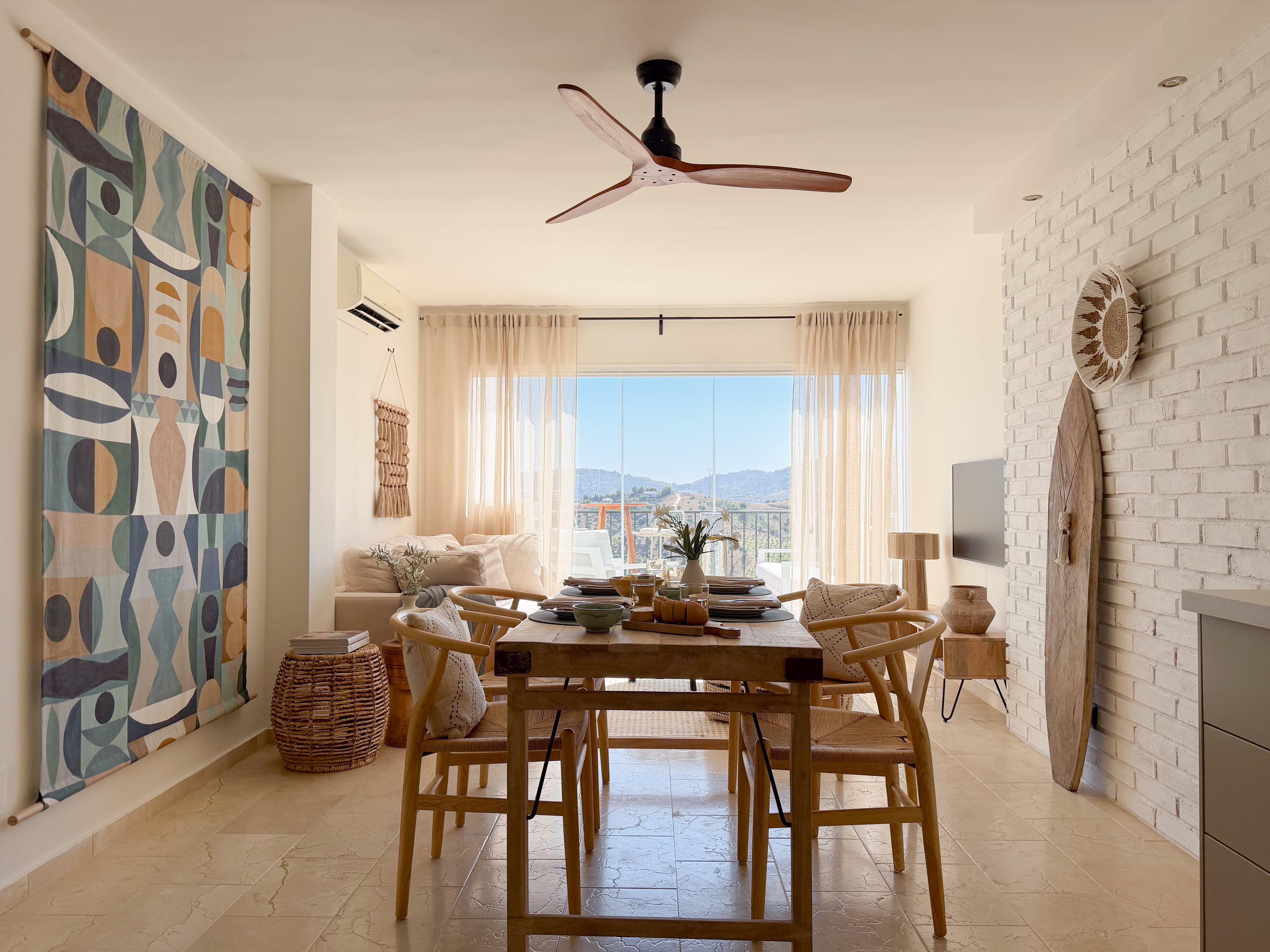 Wide view of the living and dining area with a colorful tapestry, surfboard wall art, and mountain views through the terrace doors