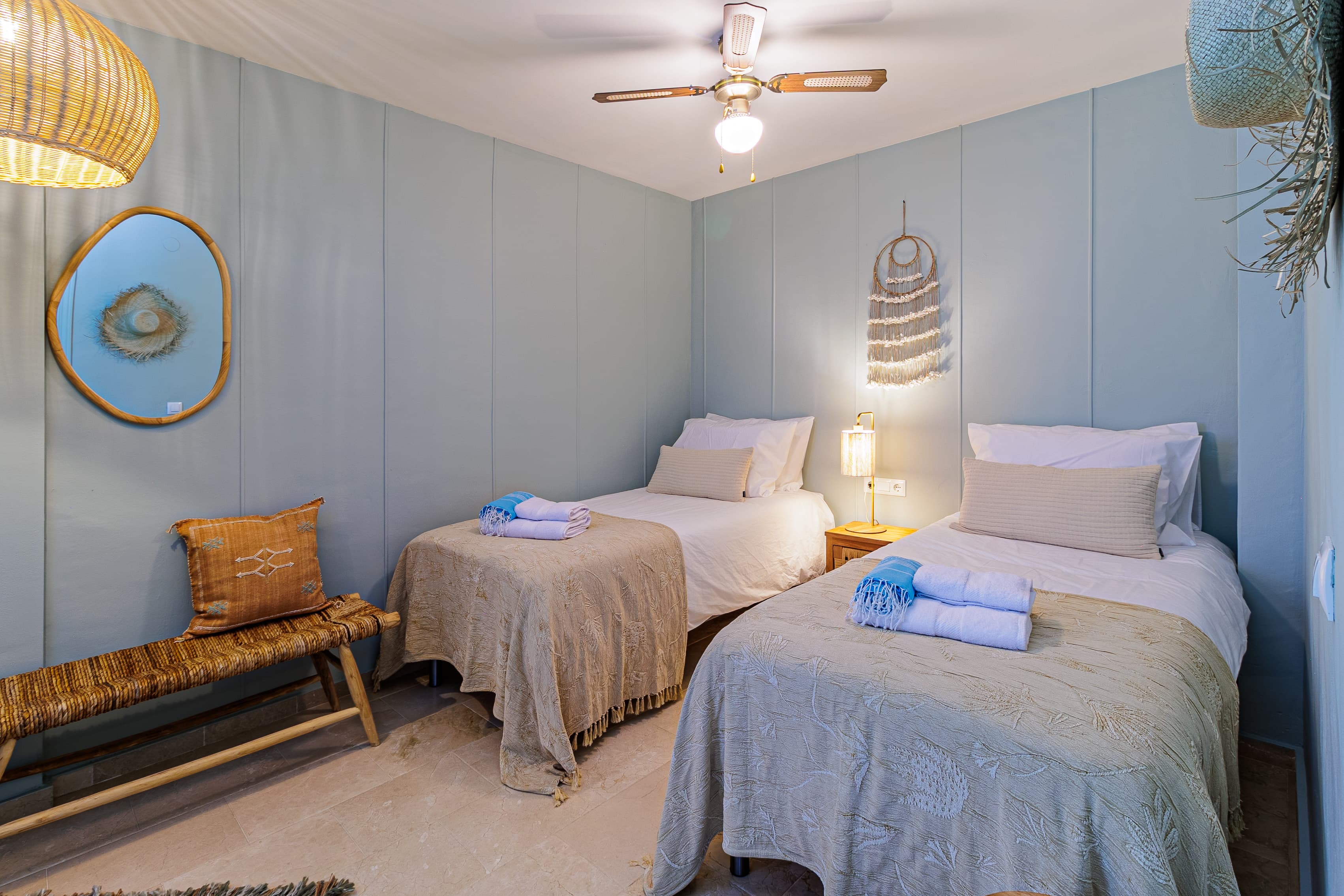 Twin bedroom with pale blue paneled walls, a woven shell wall hanging, and a rattan pendant lamp